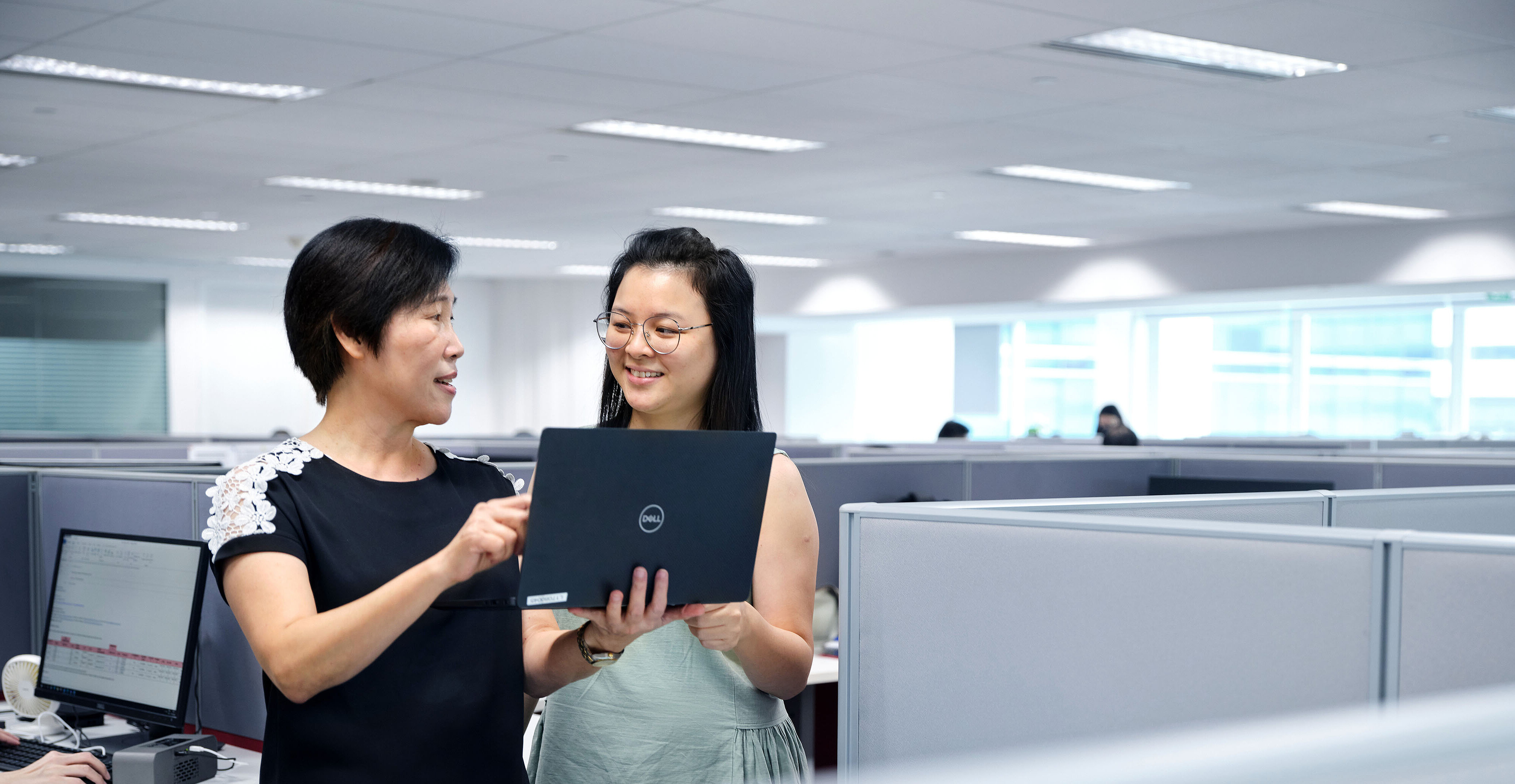 Two office colleagues reviewing content on a laptop in a modern workspace with cubicles and computer workstations
