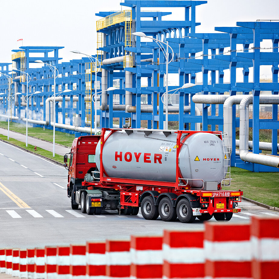 红色卡车运载 HOYER 化学罐式集装箱穿过工业厂区，蓝色钢结构及管道背景 