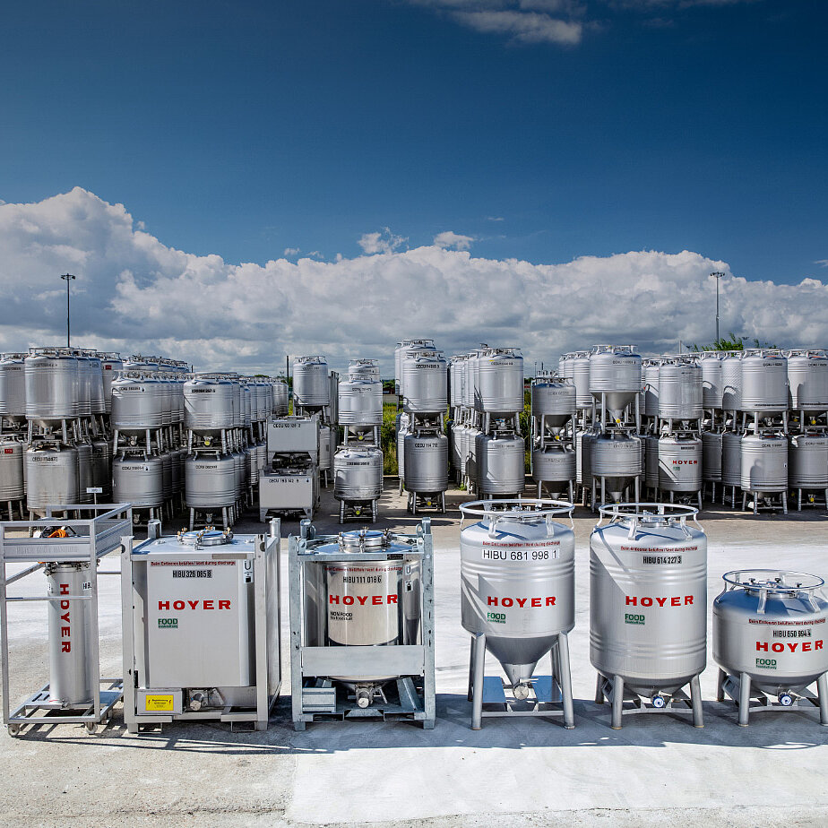 室外储存场，堆放各种尺寸的 HOYER IBC 容器，放置在金属货架上，天空部分多云