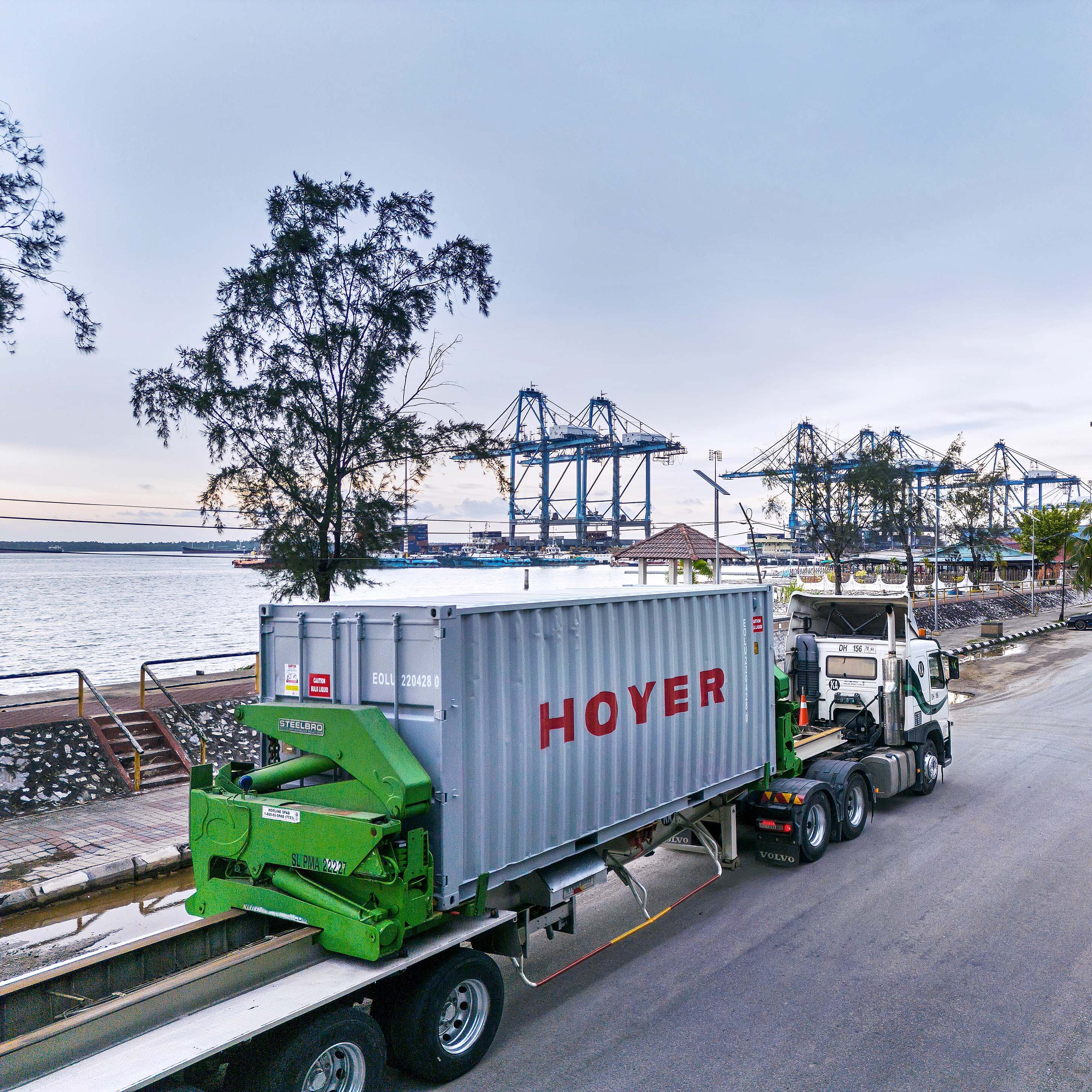 Truck transporting HOYER flexitank container on green chassis near port with cranes and water in the background