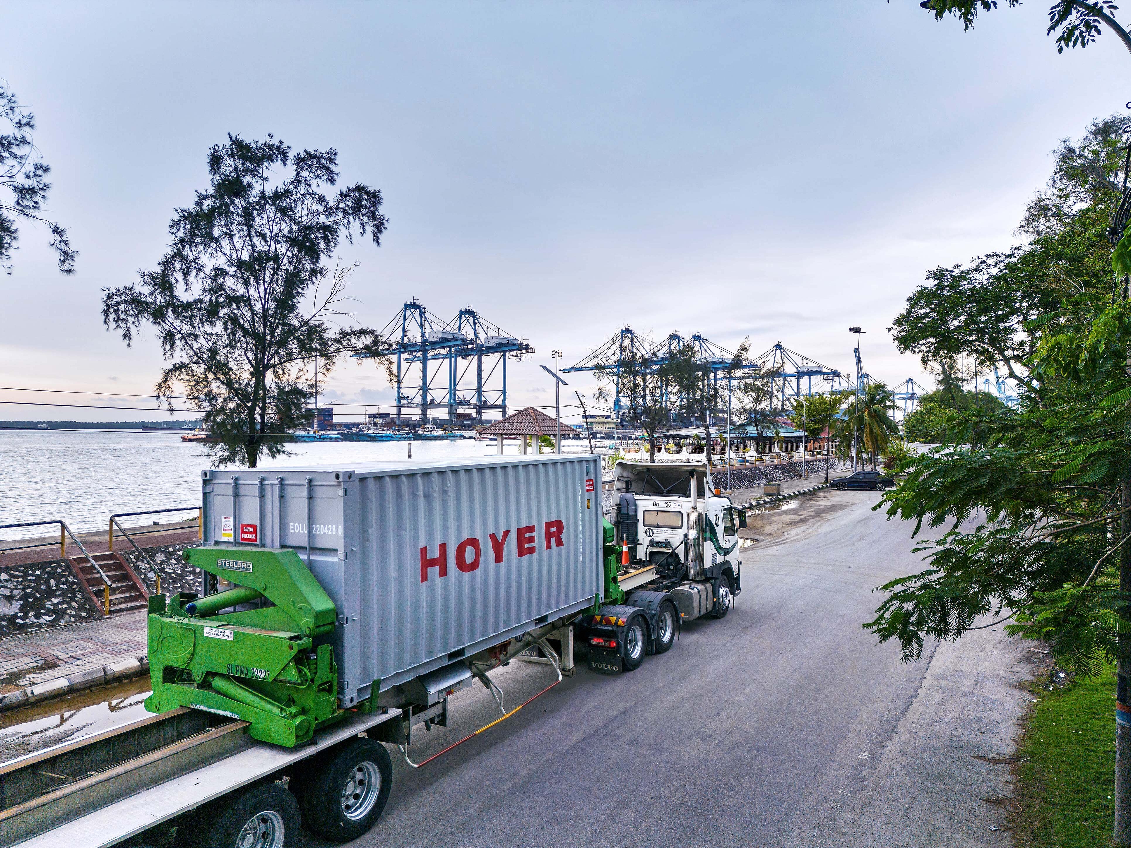 HOYER 液袋 集装箱 货车 港口 路边 卡车载着绿色底盘的 HOYER 集装箱液袋集装箱行驶于港口旁,背景可见起重机和水面