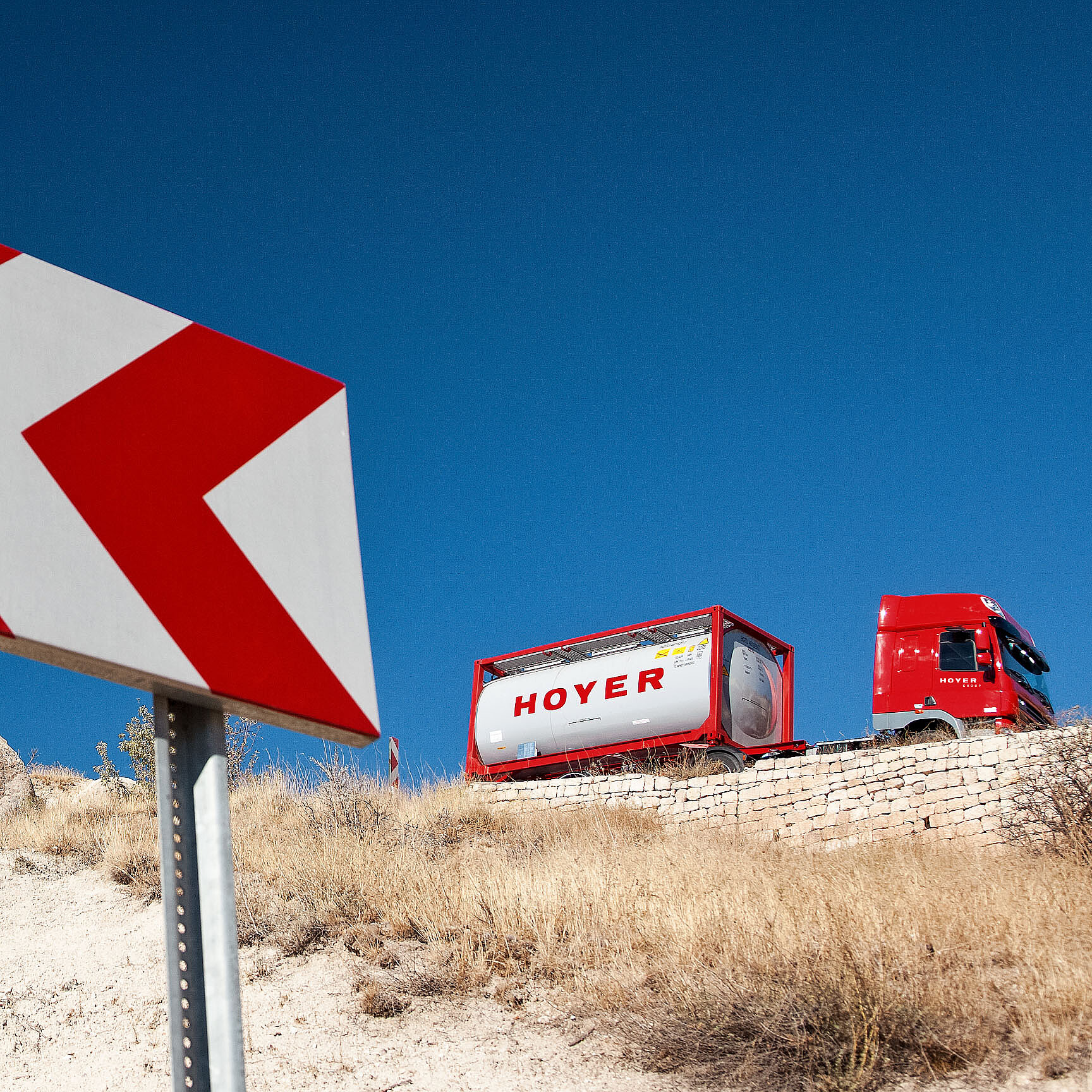 一辆红色卡车在干燥的道路上运输 HOYER 化工罐式集装箱，路旁设有警示箭头标识