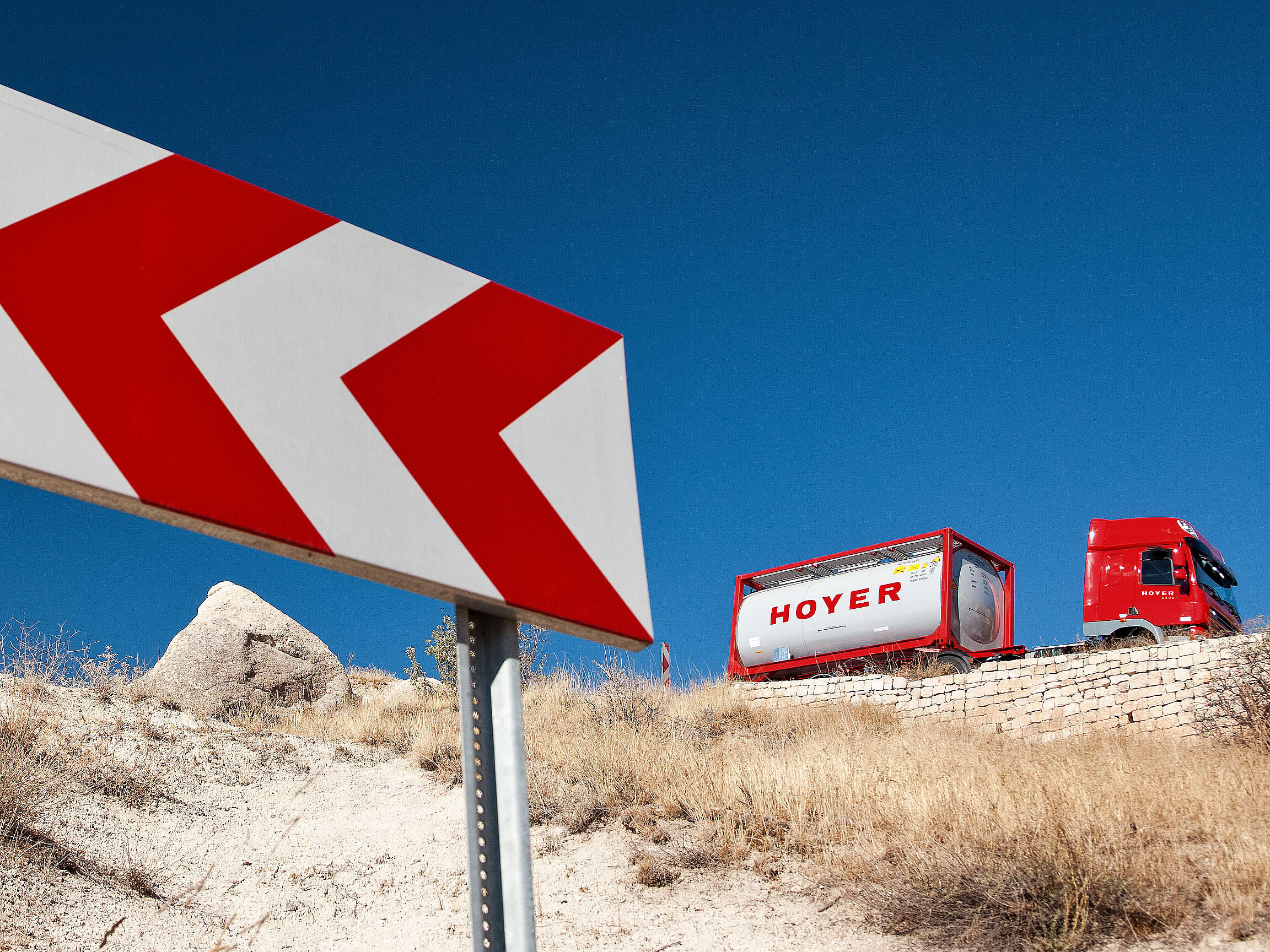 红色卡车在道路上运输 HOYER 化工罐式集装箱，路旁有指示箭头标志，干燥景观为背景
