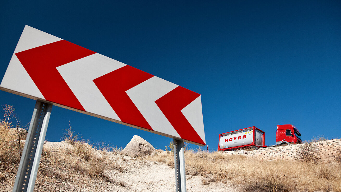 Red truck transporting a HOYER chemical tank container on a road with chevron sign and dry landscape