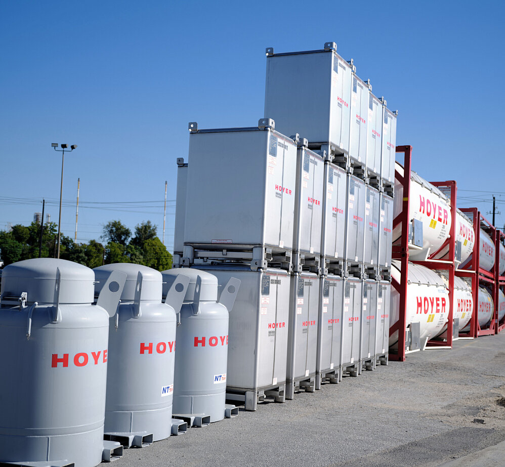 露天堆场内堆放的 IBC 容器和 HOYER 罐式集装箱，背景有树木、电线及蓝天