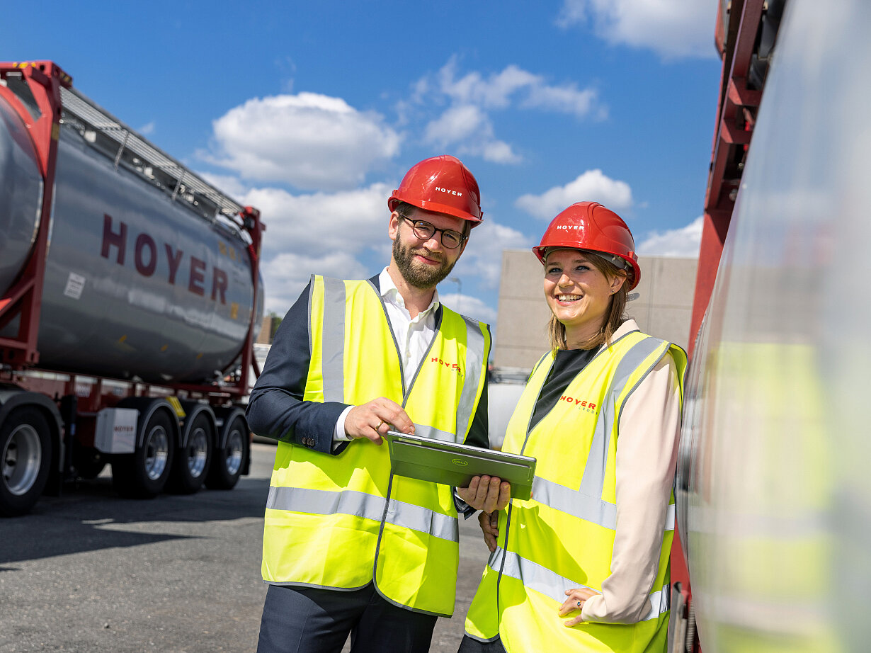 HOYER 员工 化学 罐式集装箱 高可见性 安全帽 平板电脑 两名员工穿着高可视背心和红色安全帽,站在 HOYER 化学罐式集装箱旁,共同查看平板