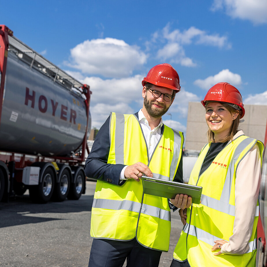 HOYER 员工 化学 罐式集装箱 高可见性 安全帽 平板电脑 两名员工穿着高可视背心和红色安全帽,站在 HOYER 化学罐式集装箱旁,共同查看平板