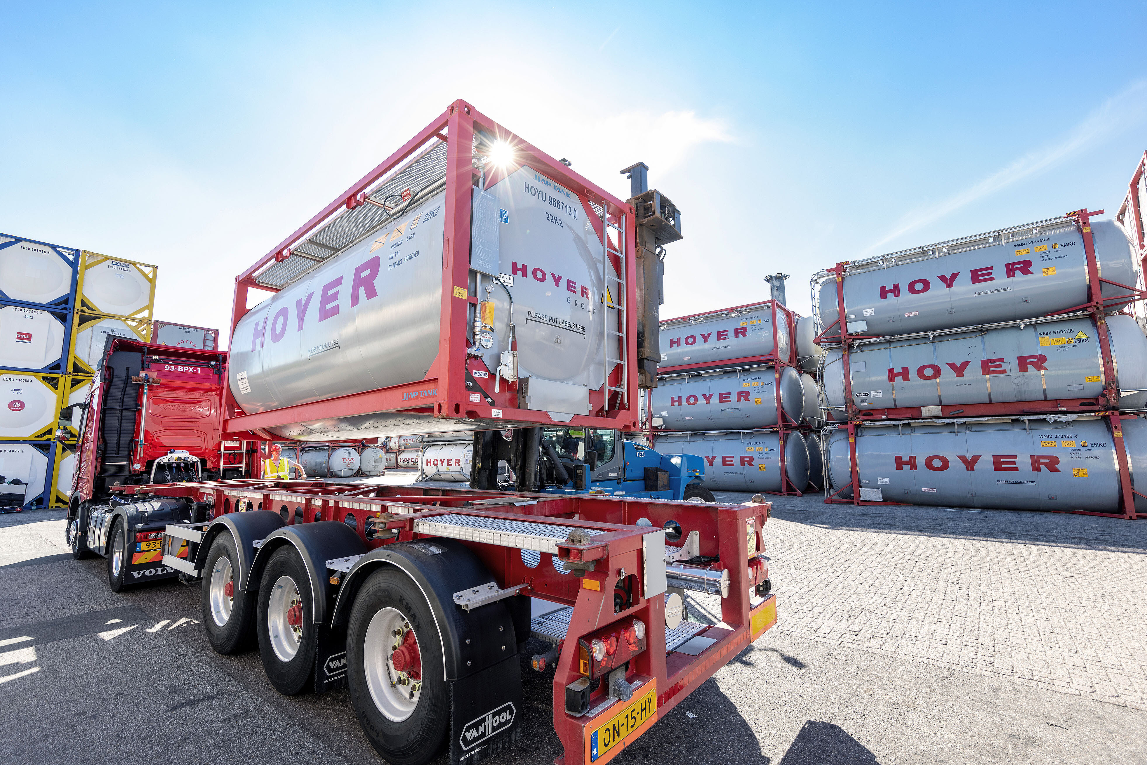 Forklift loading HOYER chemical tank container onto truck chassis at depot site with stacked containers in the background