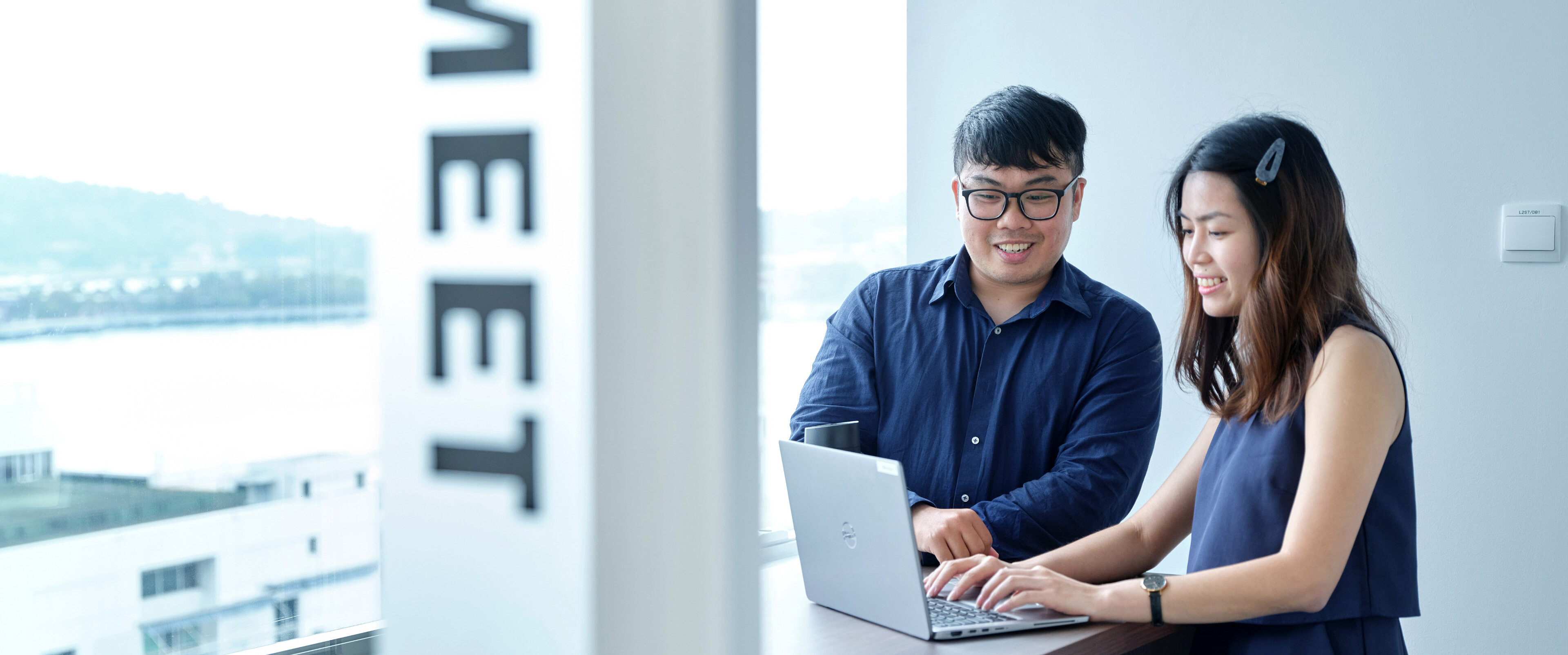 HOYER 同事 合作 笔记本 办公室 两位同事在大窗户旁协作使用笔记本电脑,窗外可见城市及水景,现代工作空间被自然光充盈