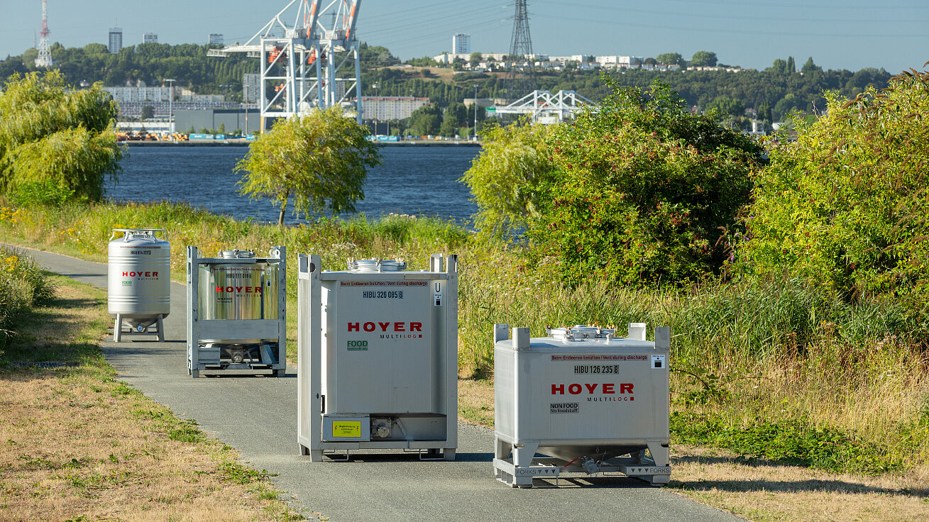 HOYER-labeled IBCs positioned along pathway near water, with cranes, industrial buildings, and greenery in the background