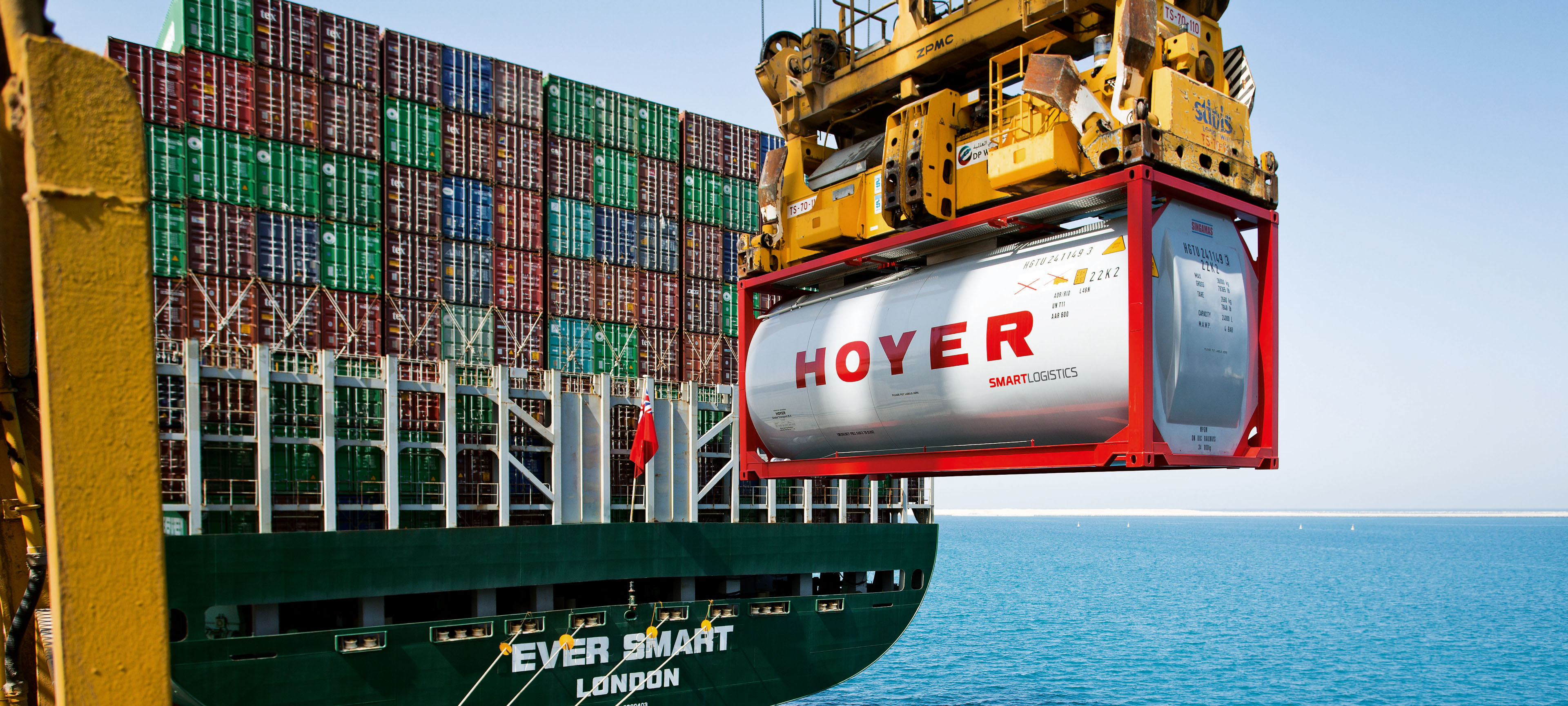 HOYER 化学罐式集装箱正在起重机吊装上货船，背景为堆叠的容器和水面