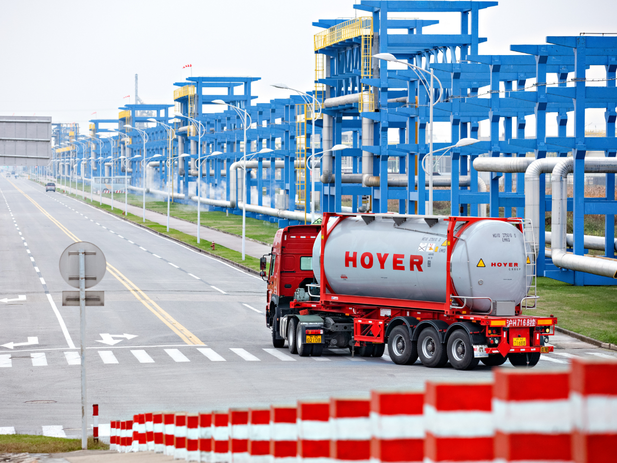 红色卡车运载 HOYER 化学罐式集装箱穿过工业厂区，蓝色钢结构及管道背景 