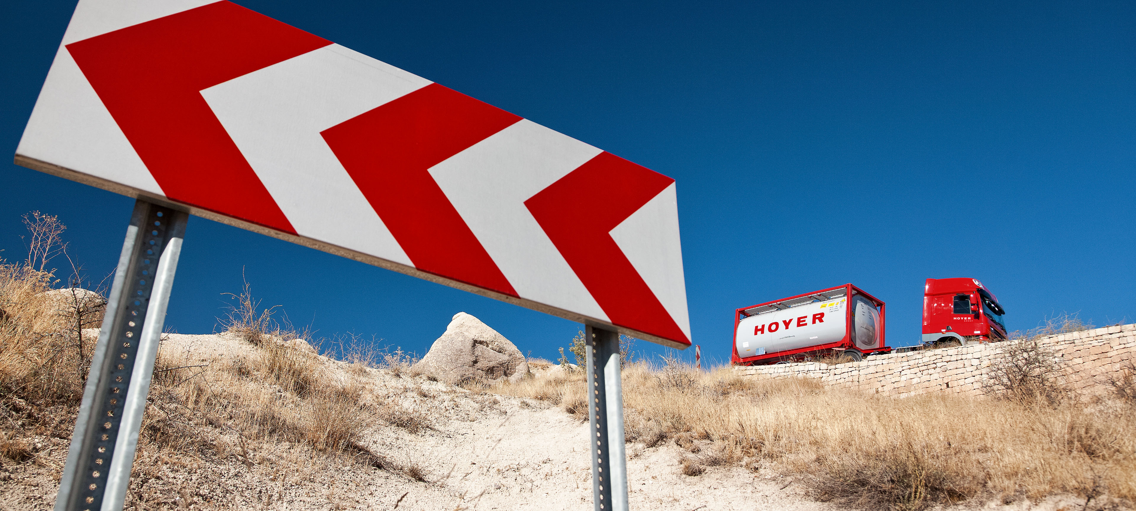 红色卡车在道路上运输 HOYER 化工罐式集装箱，路旁有指示箭头标志，干燥景观为背景