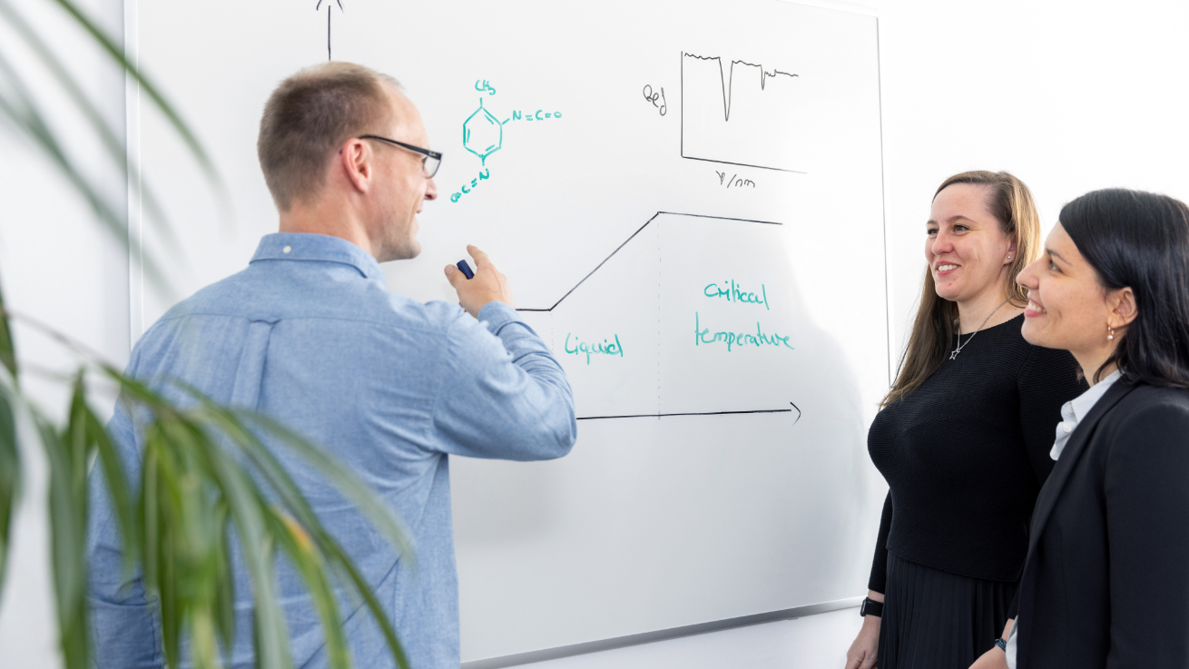Three people standing in front of a whiteboard with chemical structures, phase diagrams and measurement graphics, one person writes with a marker