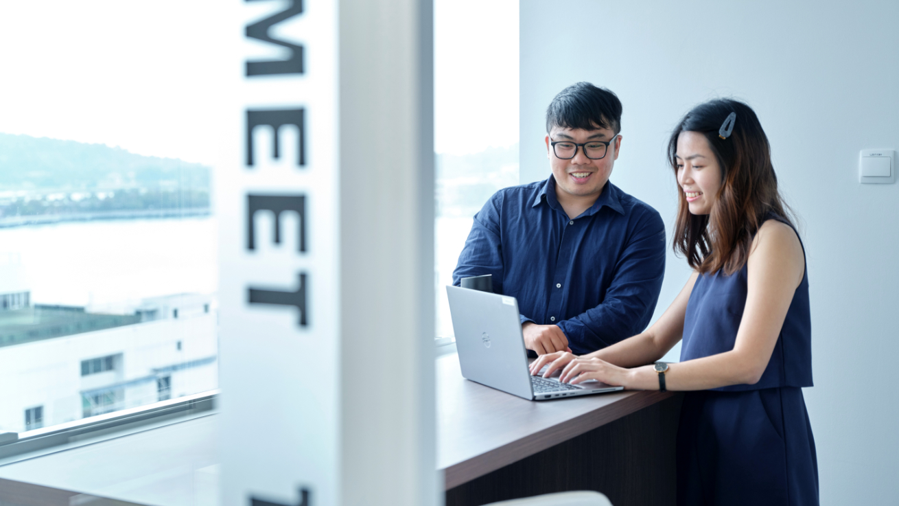 两位同事在大型窗户旁的笔记本上协作，现代办公区自然光充足，可见城市及水景