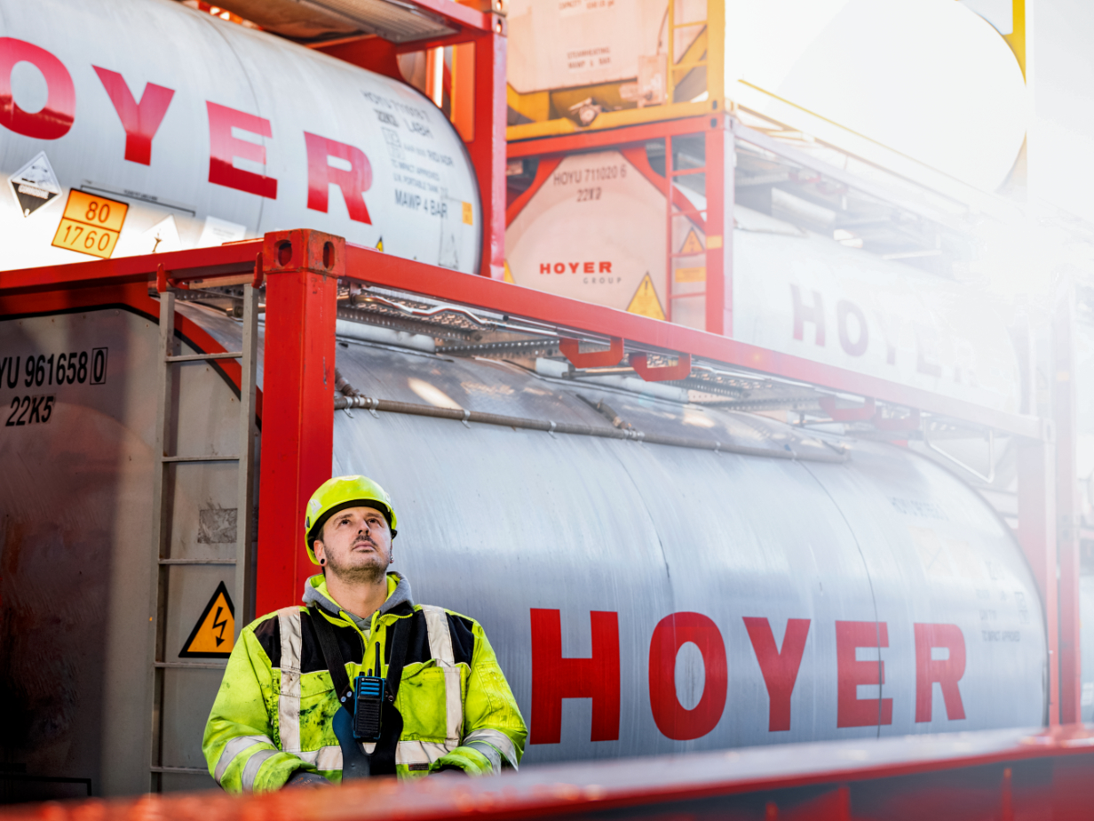 HOYER employee in high-visibility gear at dangerous goods terminal with stacked tank containers and warning labels