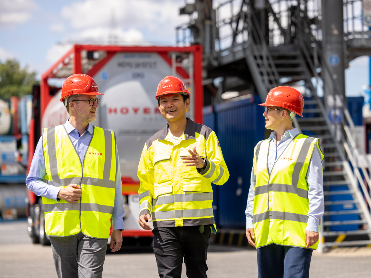 三名员工穿着高可见度背心和红色安全帽，站在 HOYER 化工罐式集装箱前，背景为工业设施