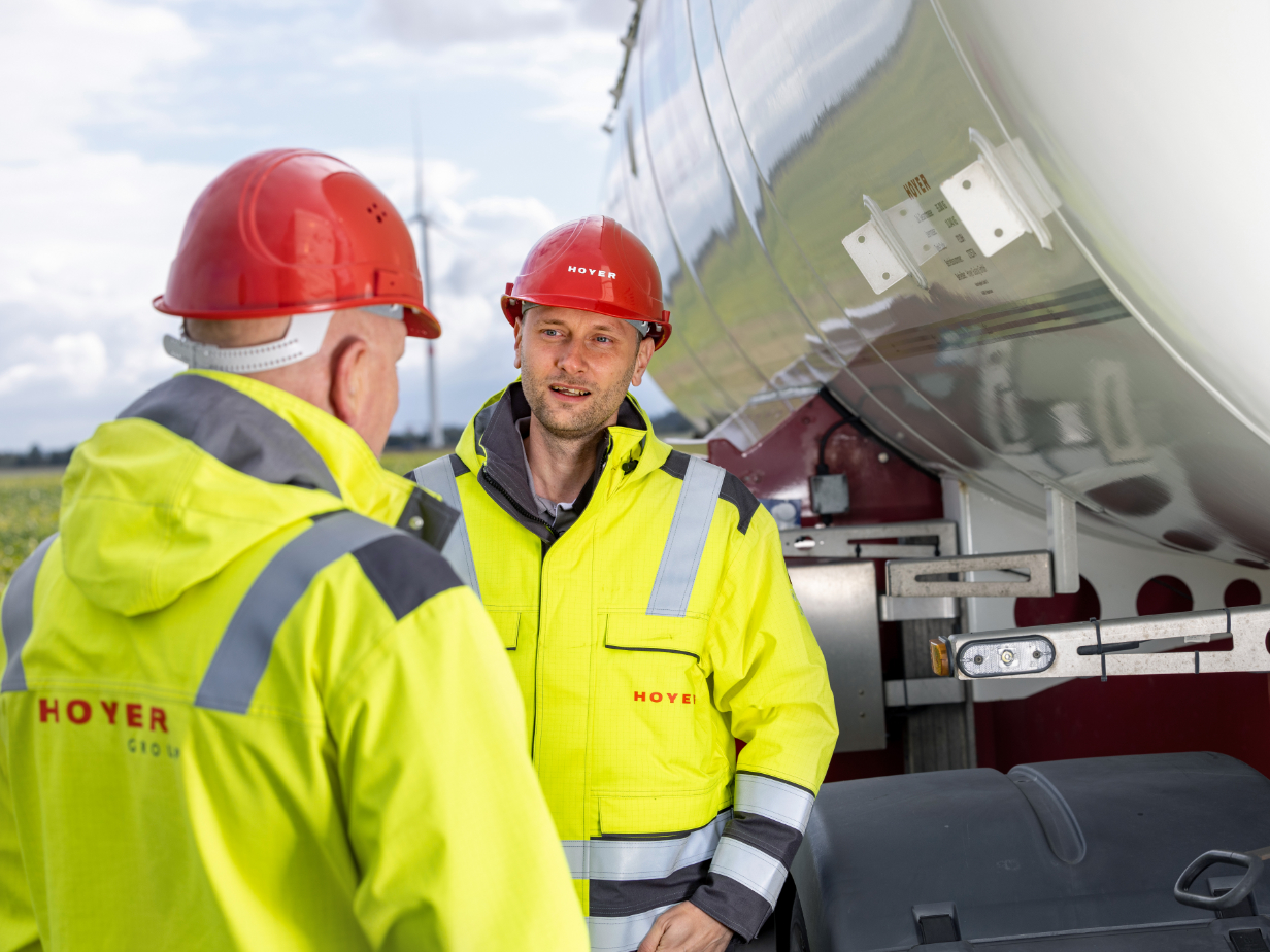 两名 HOYER 员工穿高可见度外套及红色安全帽，站在白色气体罐式集装箱车前