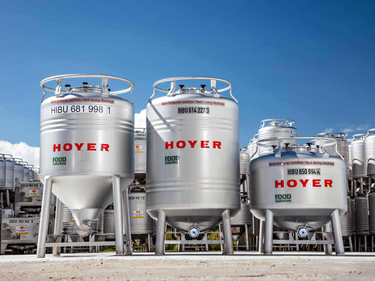 三只带编号和绿色 “FOOD” 标签的不锈钢 HOYER 食品 IBC ，户外堆放在蓝天下