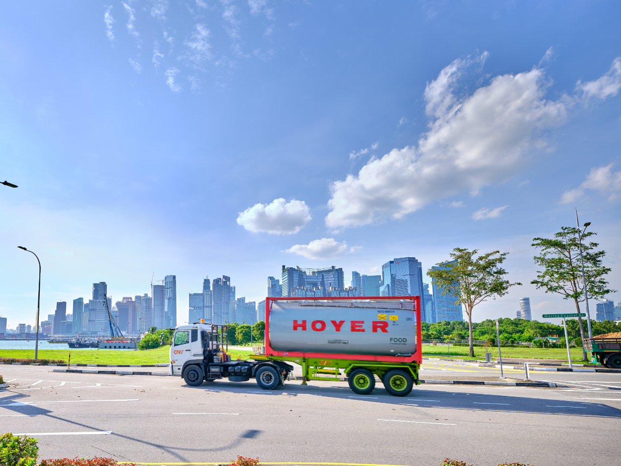 HOYER 食品罐式集装箱卡车行驶在道路上,背景为城市天际线、树木及蓝天白云 HOYER 食品罐式集装箱卡车行驶在道路上,背景为城市天际线、树木及蓝天白云