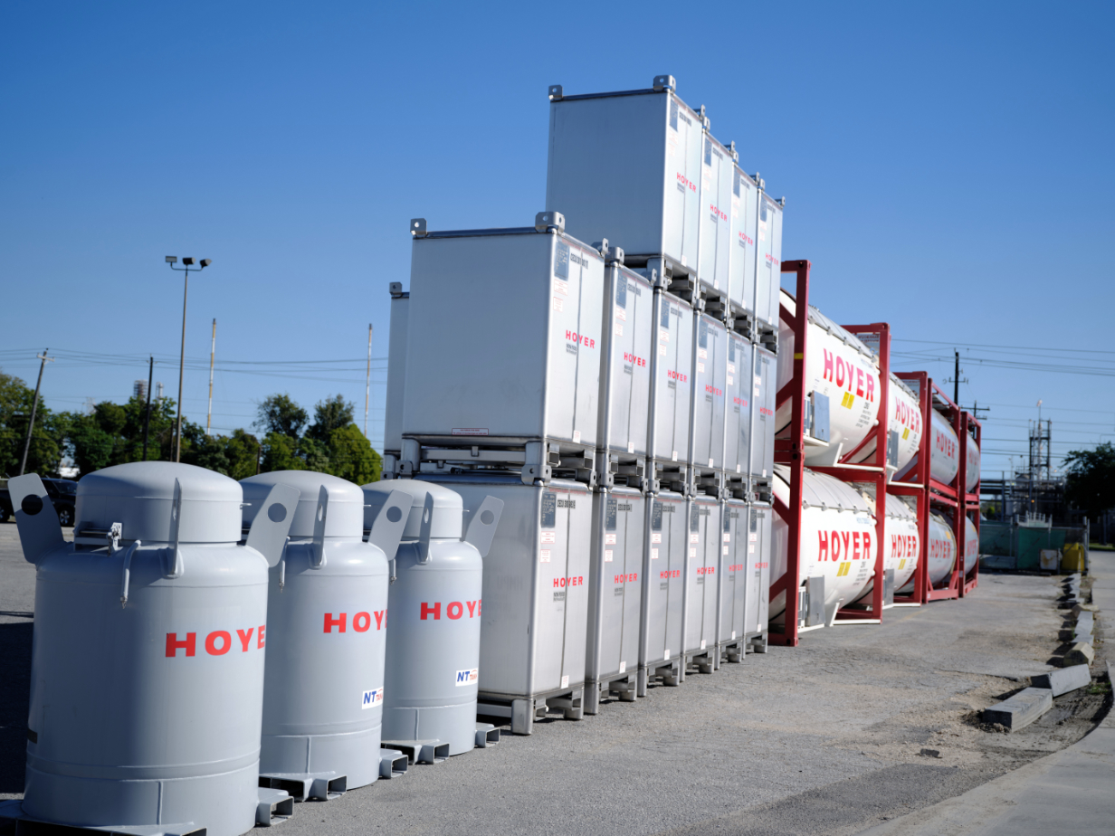 HOYER IBCs tank containers depot Stacked IBC totes and HOYER tank containers in outdoor depot area with trees, power lines, and blue sky in the background