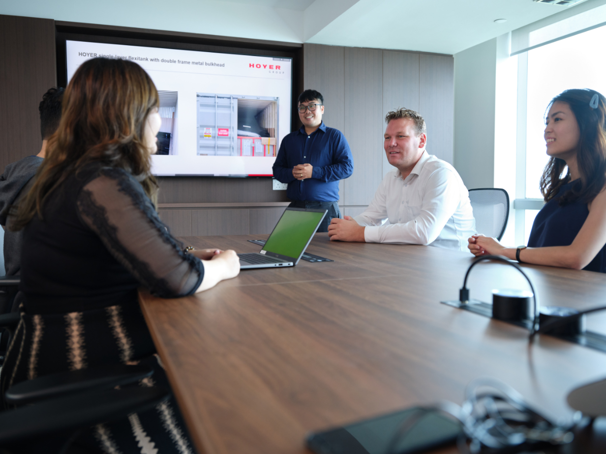 HOYER 团队在会议室内，演示者在屏幕上展示 flexitank 幻灯片，同事围坐会议桌