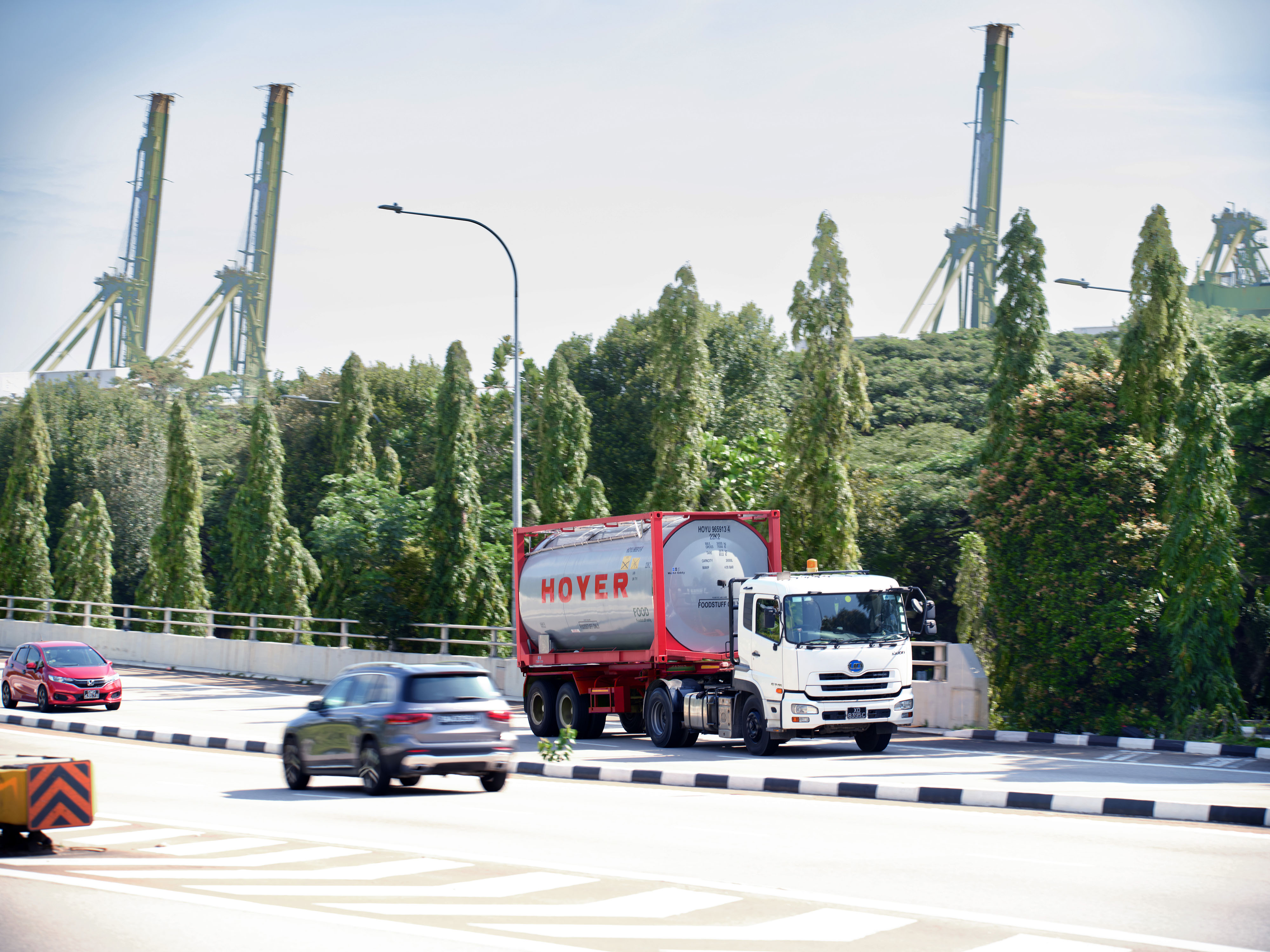 一辆装载HOYER食品罐式集装箱的白色卡车行驶在林荫大道上，背景中有两辆汽车